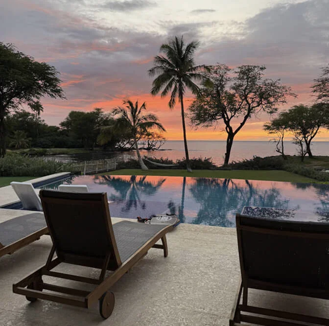 A photo of a sunset over the sea seen from a pool terrace, with palm trees, a still pool and wooden loungers in the foreground.